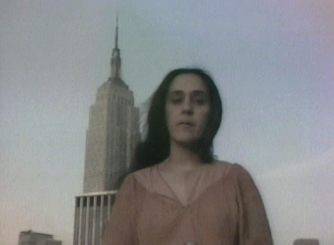 Una persona con cabello largo y ropa color marrón está frente a un edificio alto con una aguja en la cima bajo un cielo despejado.

Description generated by AI