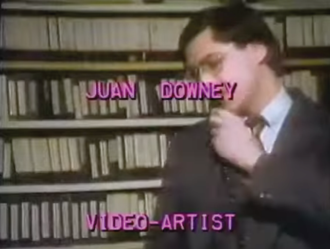 Una persona con gafas y traje sostiene un micrófono frente a estantes llenos de cintas de video con texto en pantalla que dice Juan Downey video-artist.

Description generated by AI