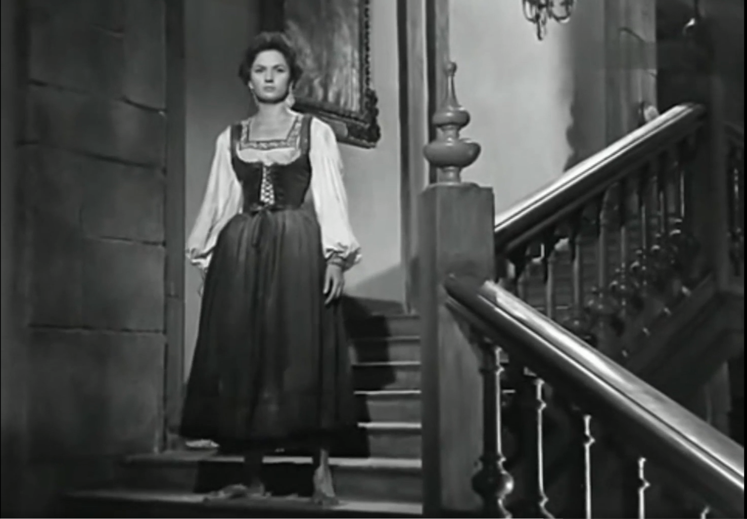 Una persona con vestido tradicional de estilo antiguo está de pie en la parte inferior de una escalera interior junto a una barandilla de madera.

Description generated by AI