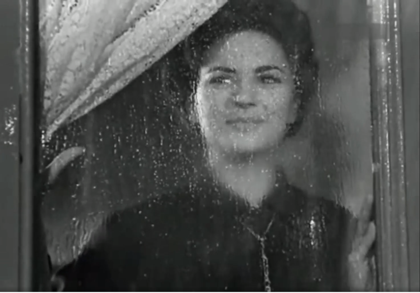 Una persona con cabello rizado y ropa oscura mira a través de una ventana con gotas de lluvia mientras sostiene una cortina blanca.

Description generated by AI