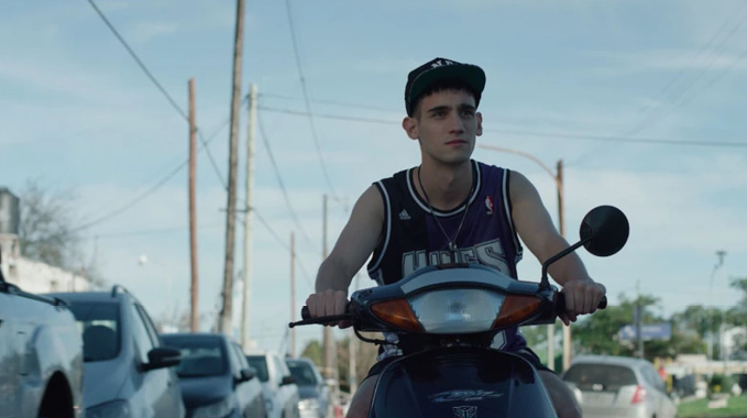 Una persona con gorra y camiseta sin mangas conduce una motocicleta en una calle con varios autos estacionados a los lados bajo un cielo despejado.

Description generated by AI