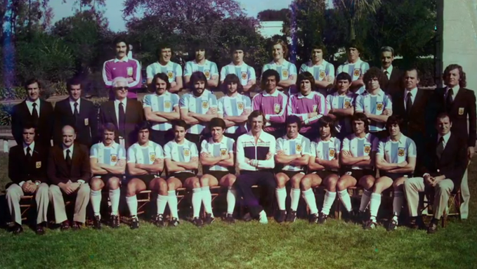Grupo grande de personas posando al aire libre en tres filas con uniformes deportivos y trajes formales, en un ambiente de equipo deportivo.

Description generated by AI
