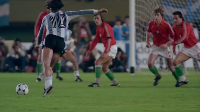 Un jugador con camiseta a rayas blancas y celestes y pantalones cortos negros está a punto de patear un balón de fútbol mientras varios jugadores con camiseta roja y pantalones cortos blancos lo observan en un campo de fútbol.

Description generated by AI