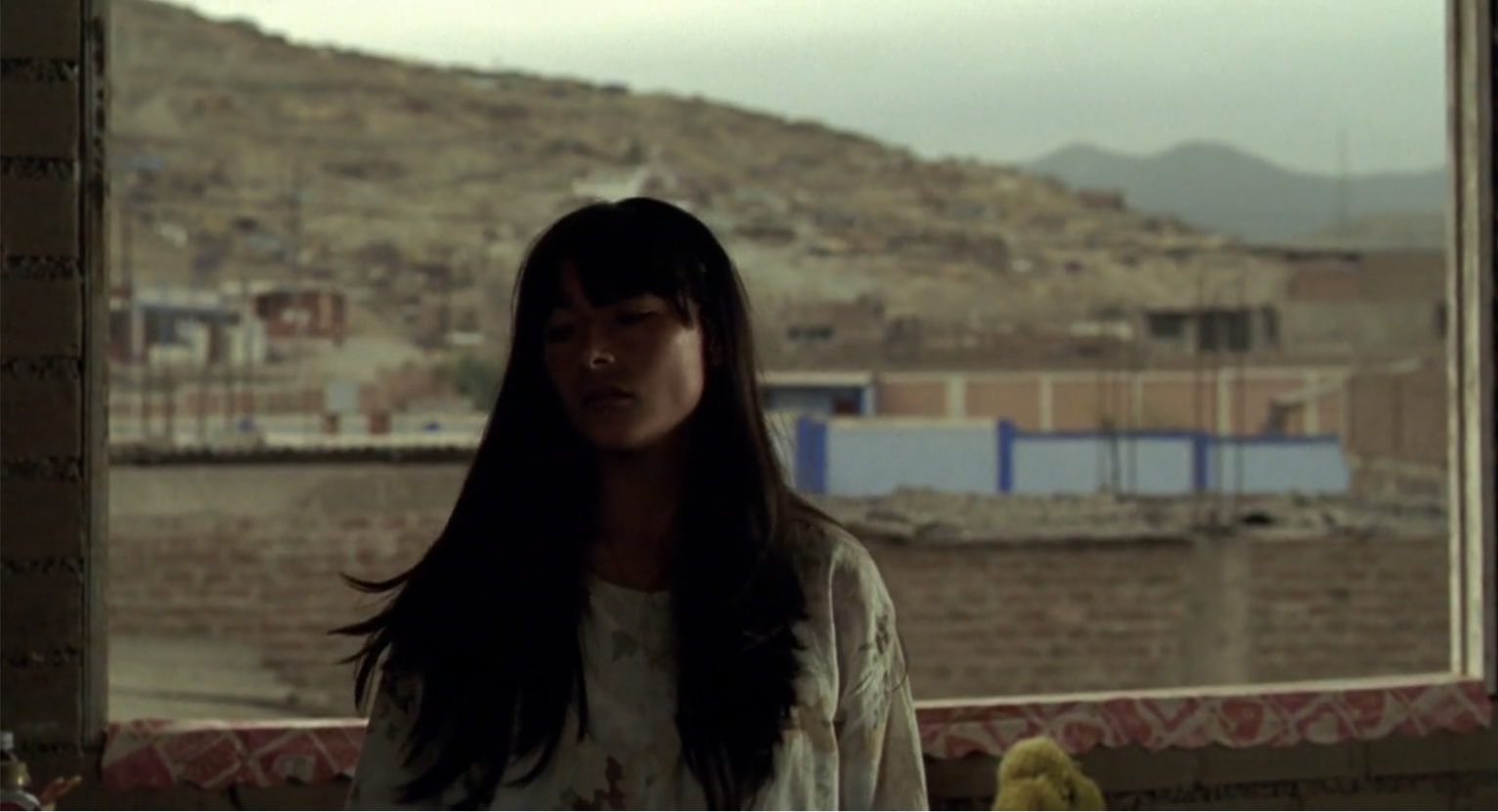 Una persona con cabello largo y oscuro está sentada frente a una ventana grande que muestra un paisaje urbano con colinas y edificios al fondo.

Description generated by AI