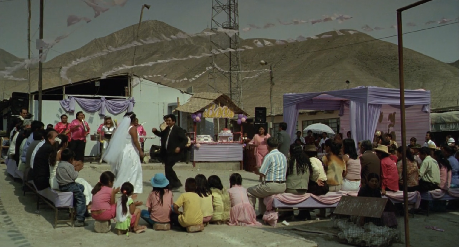 Un grupo de personas sentadas y niños en el suelo observan a dos adultos vestidos de novios bailando en una ceremonia al aire libre con montañas de fondo.

Description generated by AI